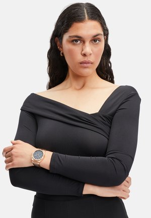 Haut noir ajusté avec un design dénudé sur les épaules, manches longues et texture de tissu drapé. Le mannequin porte une montre en or rose et de simples boucles d'oreilles.