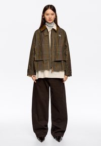 Brązowa krótka kurtka w kratę z zamkiem, z kontrastowym zamszowym kołnierzem i dwoma przednimi kieszeniami, noszona na jasnej bluzce i ciemnych szerokich spodniach.