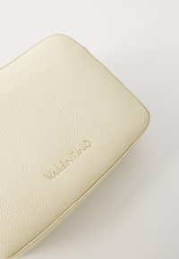 Krémová kožená kabelka na ruku s texturou kamínků, zaoblenými rohy a vyraženým logem "Valentino" ve zlatě. Minimalistický design a neutrální barva.