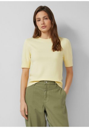 Młoda kobieta o długich brązowych włosach, ubrana w bladożółty sweter z krótkim rękawem oraz oliwkowe spodnie z wysokim stanem, stojąca z jedną ręką w kieszeni.