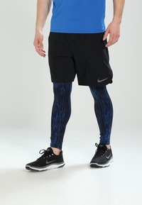 Svarta shorts, blå åtsittande leggings med geometriskt mönster och svarta träningsskor. Materialet verkar andas och vara flexibelt.