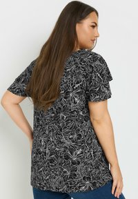 Top nero con stampa floreale bianca, vestibilità rilassata, maniche corte, orlo curvo, con una texture liscia. Abbinato a jeans blu.