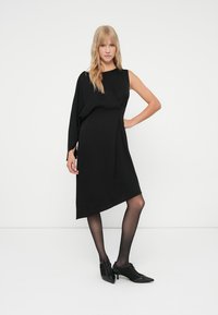 Sieviete ar blondiem matiem, tērpusies asimetriskā melnā kleitā, caurspīdīgās melnās zeķbiksēs un melnās augstpapēžu šņorētās kurpēs, stāv uz vienkārša balta fona.