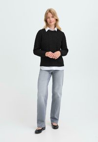 Maglione nero a maglia con collo rotondo, indossato sopra una camicia bianca con colletto, abbinato a pantaloni grigio chiaro e ballerine nere.
