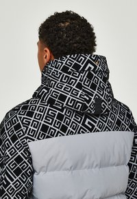 Giubbotto imbottito con cappuccio in bianco e nero a motivo, caratterizzato da disegni geometrici, sezione inferiore grigia e una finitura in tessuto testurizzato.