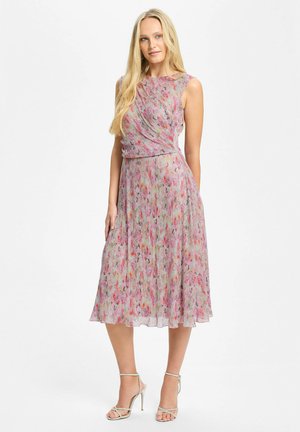 Blondynka stoi, ubrana w bezrękawnikową, różową sukienkę midi w kwiaty o plisowanej fakturze, w połączeniu ze srebrnymi sandałami na wysokim obcasie.