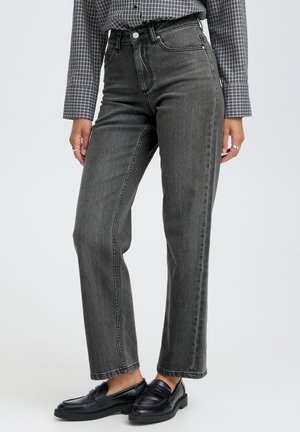 Kvinde iført grå højtaljede jeans med lige ben, en ternet skjorte stoppet ind og sorte læder loafers, stående mod en ensfarvet baggrund.