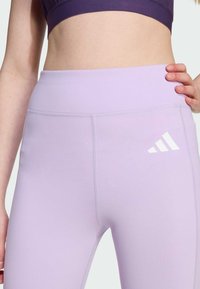 Ljust lila träningstights med bred midjeresår, slät textur och vit Adidas-logotyp på nedre vänstra sidan.