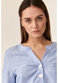Camicia di lino azzurro chiaro con scollatura a V, caratterizzata da tre bottoni rotondi al colletto. Il tessuto ha una leggera trama e lucentezza.