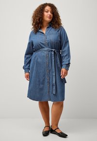 Abito chemisier in denim con maniche lunghe, chiusura frontale con bottoni e cintura in vita. Presenta una tasca sul petto e orlo fino al ginocchio in blu medio.