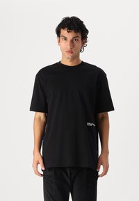 Black cotton t-shirt with a round neckline and short sleeves. Features a logo print on the left side. Simple, loose-fitting design.