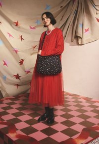 Borsa a tracolla nera con motivi di stelle colorate, maglione rosso lavorato a maglia e gonna a strati in tulle rosso. Accessori includono una collana di perle e mocassini.