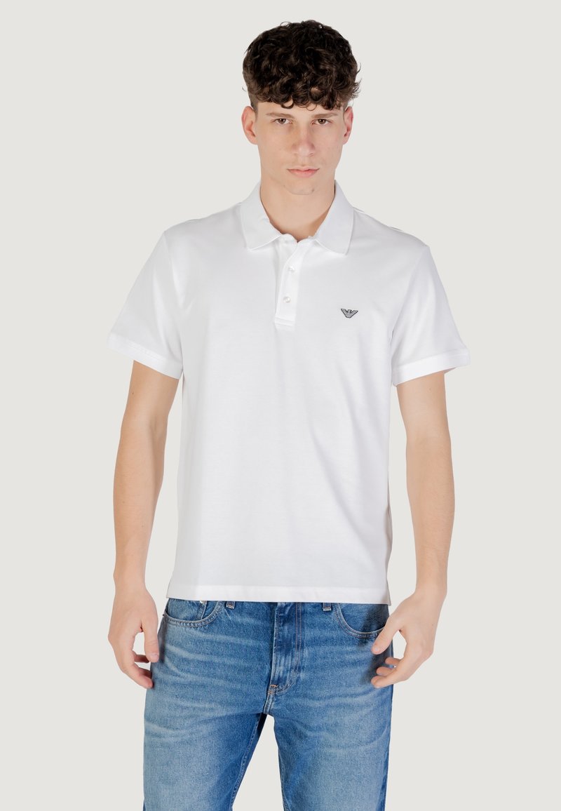 Polo shirt blanc en coton, avec un col classique, des manches courtes et un petit logo sur la poitrine gauche. Porté avec un jean bleu.