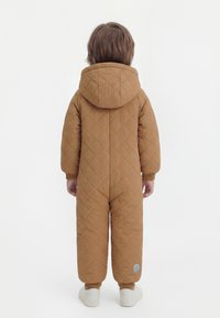 Enfant debout, vu de dos, portant une combinaison de neige marron matelassée à capuche, avec des baskets blanches, sur un fond blanc.