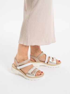 Füße in beige und metallisch silbernen Riemchensandalen mit weißen, klobigen Sohlen, kombiniert mit einem hellbeigen, gerippten Rock.