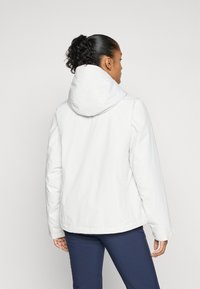 Vrouw met een donkere, krullende paardenstaart, gekleed in een witte hoodie en marineblauwe broek, gezien vanuit de achterkant tegen een effen achtergrond.