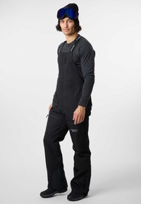 Black snow overalls with adjustable straps, front zip, and cargo pockets. Paired with a dark long-sleeve top and ski goggles.