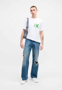 Camiseta de algodón blanca con logo verde de Calvin Klein, combinada con jeans desgastados de color azul claro con grandes desgarrones. Llevada con zapatillas blancas y un bolso gris.