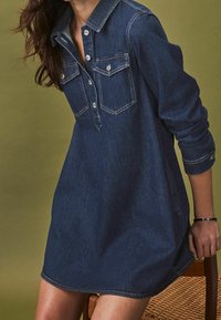 Tmavě modré denimové šaty s límcem na knoflíky, dvěma náprsními kapsami a dlouhými rukávy. Obsahují stříbrné knoflíky a volnější střih.