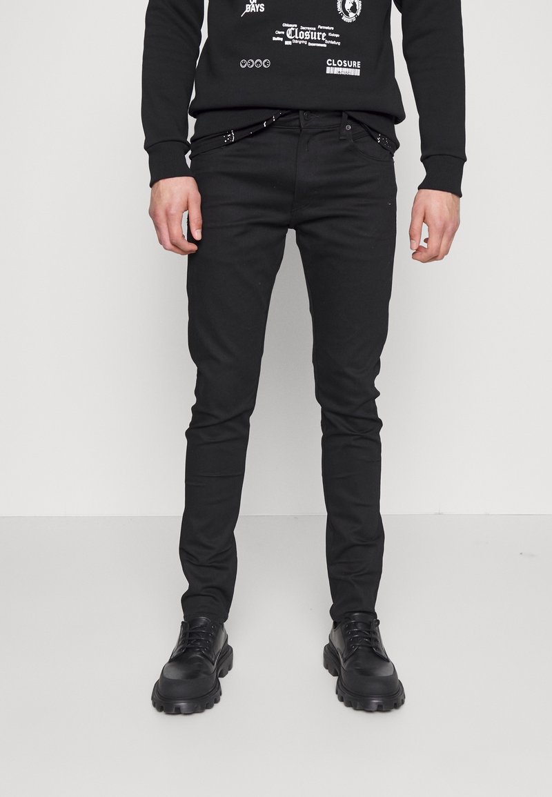 Homme portant un pantalon slim noir, des chaussures noires épaisses à lacets, et une chemise noire à manches longues avec un texte et des graphiques blancs.