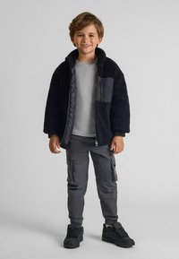 Veste en polaire noire avec zip, chemise grise en dessous, pantalon cargo gris et baskets noires. La veste présente des détails de poches contrastants.