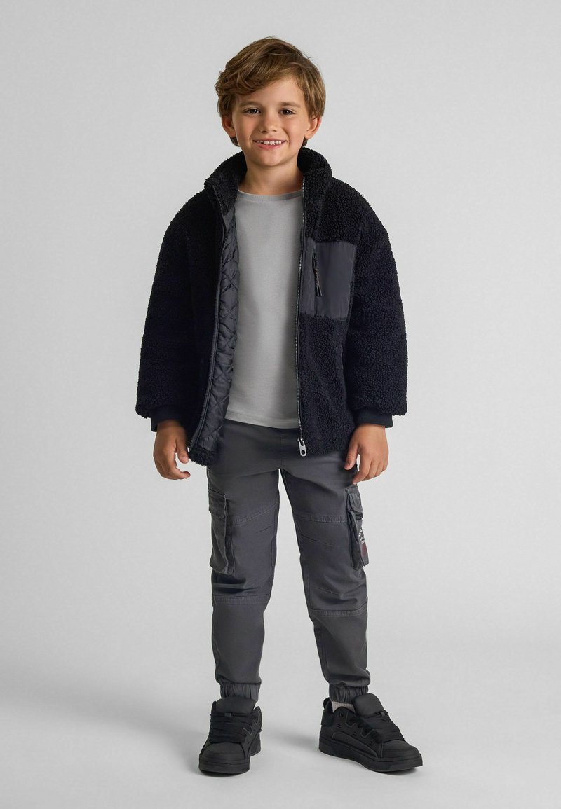Veste en polaire noire avec zip, chemise grise en dessous, pantalon cargo gris et baskets noires. La veste présente des détails de poches contrastants.