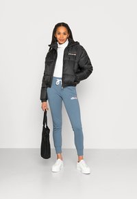 Svart pufferjakke med hette, blå joggebukser med logo, hvite sneakers og svart totebag, satt mot en ensfarget bakgrunn.