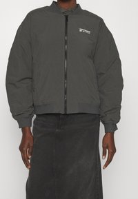 Giubbotto bomber grigio con zip frontale, polsini e orlo a coste, e piccolo logo bianco "New Balance" sul petto. Tessuto resistente e morbido.