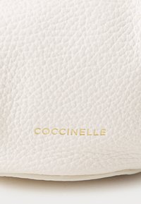 Bílá kožená kabelka s texturovaným povrchem a výrazným zlatým logem "Coccinelle". Jednoduchý design bez dalších kovových prvků.