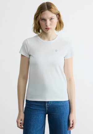 Νεαρή γυναίκα με κοντά ξανθά μαλλιά, φορώντας ένα απλό λευκό εφαρμοστό t-shirt και μπλε τζιν, στέκεται μπροστά σε ένα ανοιχτό φόντο.