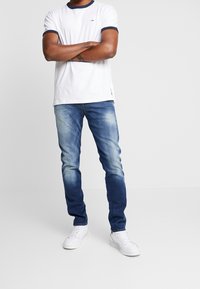 Vit t-shirt med marinblå ringerkrage, blå slitna jeans och vita sneakers. T-shirten har en liten logga på vänster bröst.