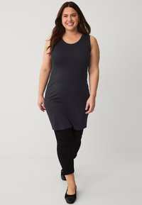 Zwarte mouwloze jurk die net boven de knie valt, gemaakt van zachte stof; gecombineerd met zwarte leggings en zwarte schoenen met een lage hak.