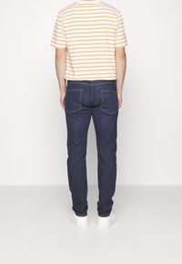 Camiseta de manga corta a rayas en blanco y naranja, combinada con jeans de mezclilla azul oscuro. Zapatillas blancas visibles, prenda mostrada desde la parte posterior.