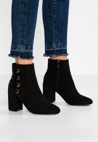 Botas de ante negras hasta el tobillo con punta afilada, cremallera lateral y diseño de cordones con ojales metálicos. Diseño de tacón cuadrado para mayor estabilidad.