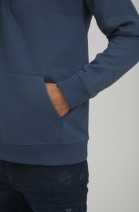 Dunkelblaues Sweatshirt aus weichem Material, mit einer Fronttasche und Rippbündchen. Getragen mit dunklen Jeans.
