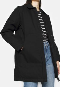 Sort jakke lavet af et struktureret stof, med krave, lynlåslukning foran og to forlommer. Båret over en stribet skjorte og blå jeans.