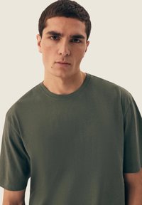Oliwkowa koszulka z okrągłym dekoltem, z krótkim rękawem, wykonana z miękkiego materiału, gładka w dotyku, z minimalnymi detalami i luźnym krojem.
