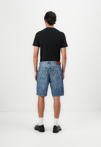 Jeansshorts mit einem gemusterten Design in Hellblau, ausgestattet mit einem braunen Lederpatch am Bund und im Five-Pocket-Stil. Schwarzes T-Shirt getragen.