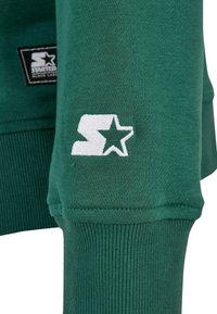 Zelena dukserica s rebrastim manžetama; sadrži bijeli izvezeni logo i oznaku. Teksturna tkanina, duži rukavi i ležeran dizajn.