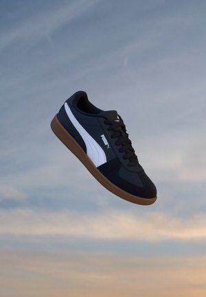 Basket Puma noir et blanc avec semelle en gomme flottant devant un ciel du soir nuageux.