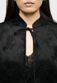 Γυναίκα που φορά ύφασμα μαύρου χρώματος με λουλουδάτο σχέδιο, δεμένο σε φιόγκο στον λαιμό, με εμφανές μαύρο δαντελένιο ύφασμα από κάτω.