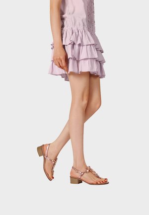 Mujer con vestido de lavanda en capas y sandalias de oro rosa con tacones bajos y anchos, de pie con una pierna cruzada delante de la otra.