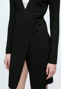 Svart ribbestrikket omslagskjole med lange ermer, med knyting i siden og høyt sliss, laget av et mykt, teksturert stoff.