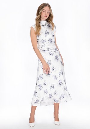 Donna con capelli biondi lunghi, che indossa un vestito bianco senza maniche con motivi floreali blu e scarpe con tacco bianco, in piedi davanti a uno sfondo bianco.