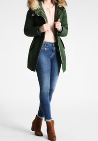 Grøn parka-jakke med en foring af pels i hætten, lynlåslukning, to frontlommer, parret med blå denimjeans og brune ankelstøvler.