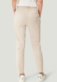 Pantalons beige, coupe slim avec une texture lisse, deux poches arrière et un design épuré. Portés avec des baskets argentées, affichant une esthétique simple.