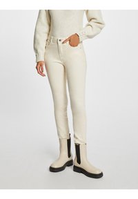 Pantalones vaqueros de denim color crema con un ajuste slim, con una cintura alta, cinco bolsillos y una textura suave. Llevados con botas de tobillo beige.
