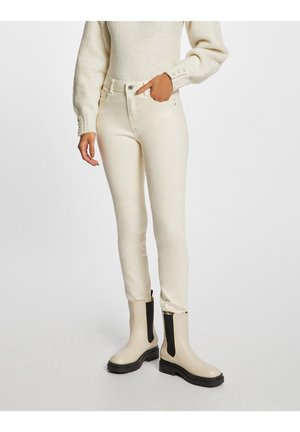 Jean en denim couleur crème avec une coupe slim, présentant une taille haute, cinq poches et une texture lisse. Porté avec des bottines beiges.