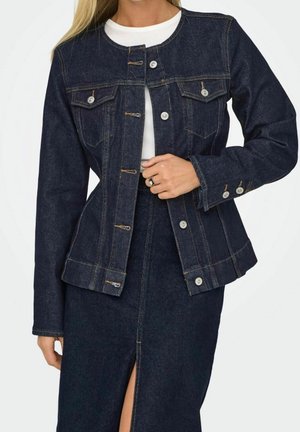 Femme portant une veste en jean bleu foncé cintrée et une jupe assortie fendue devant, tenant la veste d'une main.