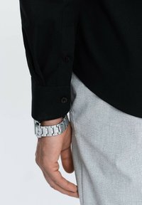 Camicia nera a maniche lunghe con polsini abbottonati, abbinata a pantaloni grigi testurizzati e un orologio argentato su una mano, che mostrano un contrasto di colori e materiali.